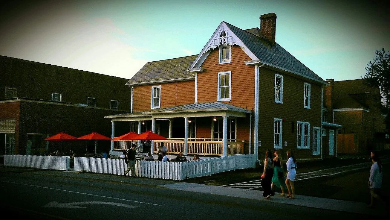 The Blacksburg Tavern at dusk: a two-storey clapboard house with white trim, a covered porch, red table umbrellas and a white picket fence, with people walking past on the sidewalk.